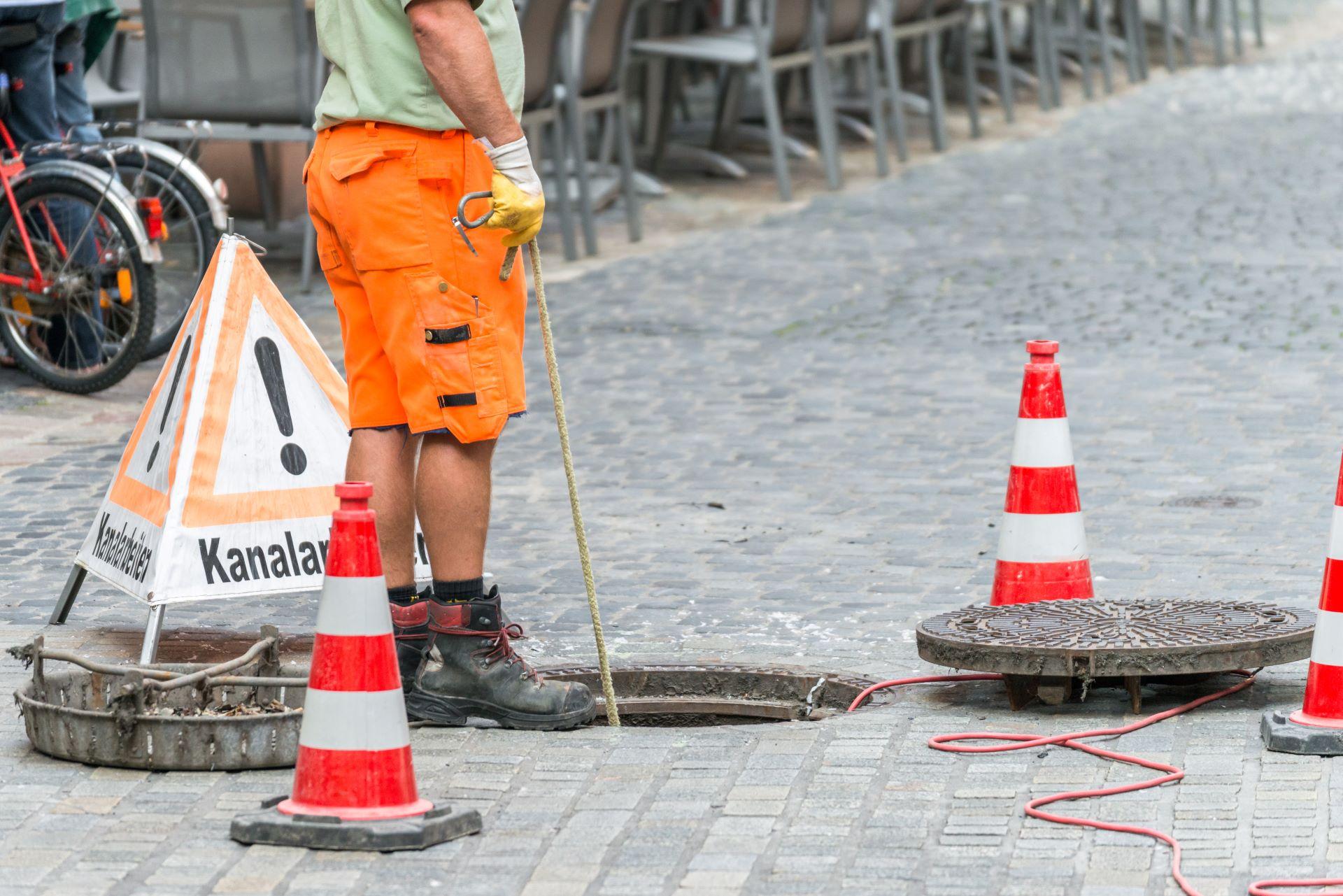 Straßenarbeiter in orangefarbener Schutzkleidung bei Kanalarbeiten, umgeben von Verkehrskegeln und einem Warnschild mit der Aufschrift "Kanalarbeiten" neben einer geöffneten Kanalabdeckung.